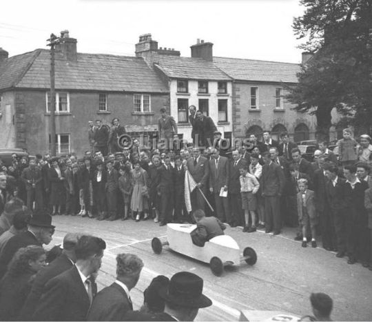 black and white photograph of finish line for soap box race showing car approaching finish line with spectators watching.